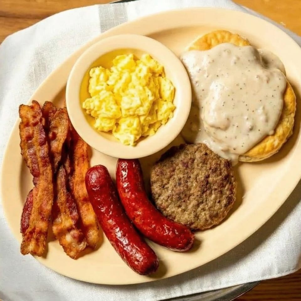 Hearty breakfast plate with scrambled eggs, bacon, sausages, sausage patty, and biscuits with gravy.