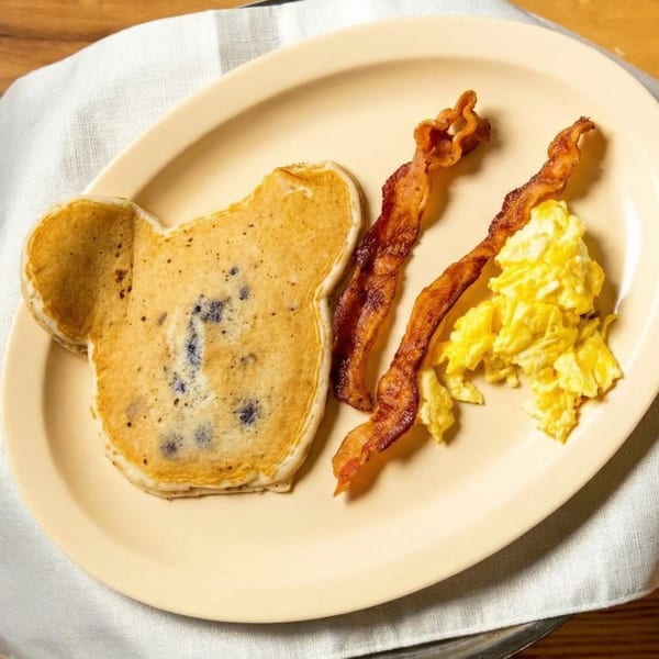 Bear-shaped blueberry pancake, crispy bacon, and scrambled eggs on a beige plate.