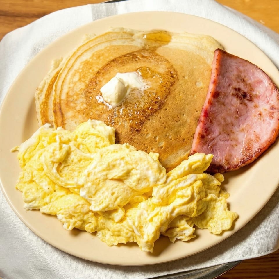 A breakfast plate with pancakes, scrambled eggs, and a slice of ham on a light yellow plate.